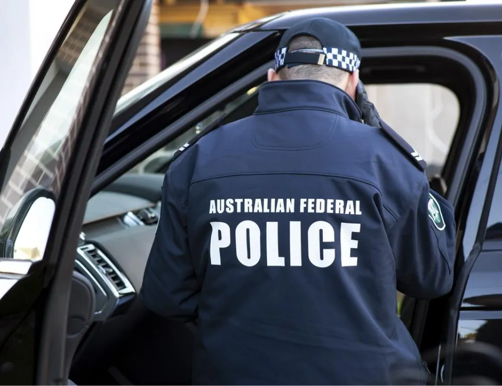 A police officer wearing a jacket with "Australian Federal Police" stands beside a vehicle.