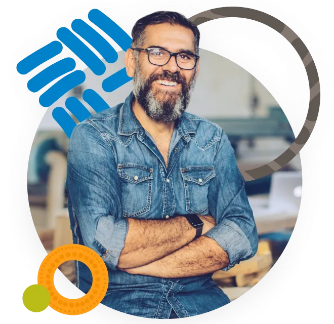 Smiling man with a beard, wearing a denim shirt, sitting with arms crossed.