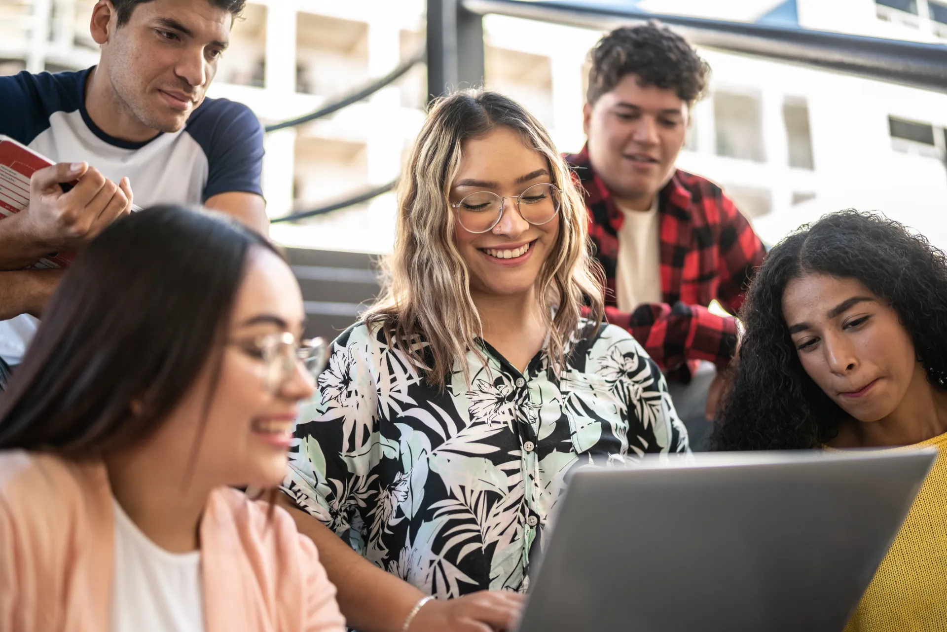 Five young adults gather around a laptop, smiling and collaborating on a project.