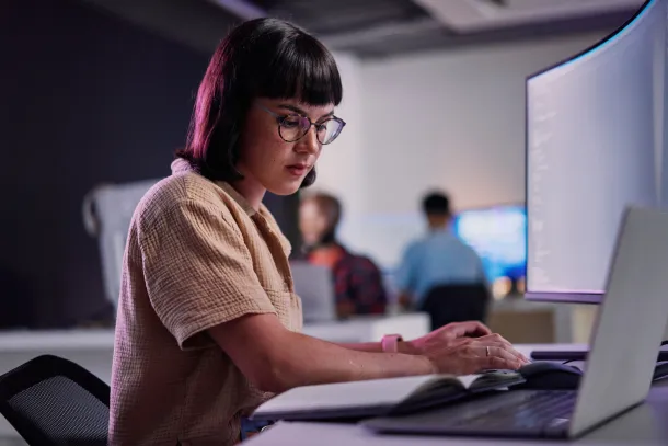 A woman with short hair and glasses works intently at a computer in a modern office setting.