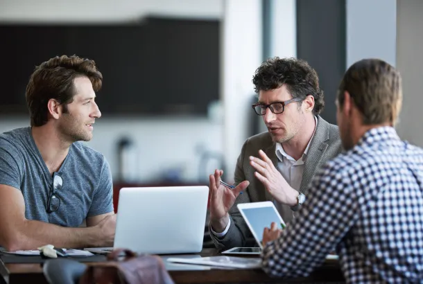 Three men engage in a discussion at a table with laptops and a tablet.