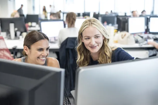 Two women smile while looking at a computer screen in a modern office environment.