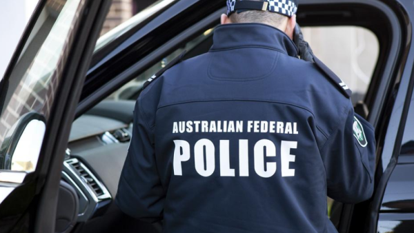 A police officer wearing a jacket with "Australian Federal Police" stands beside a vehicle.