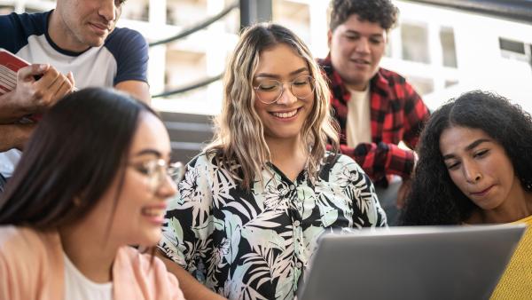 Five young adults gather around a laptop, smiling and collaborating on a project.